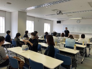 青年交流会、教室方式で、佐藤さんの説明を聞いている14名の参加者とボランティアのみなさん