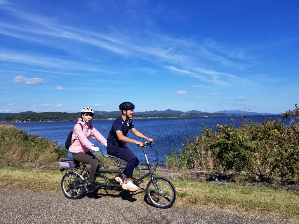 宍道湖を経背景にタンデム自転車に乗って走るペアの写真（行き）